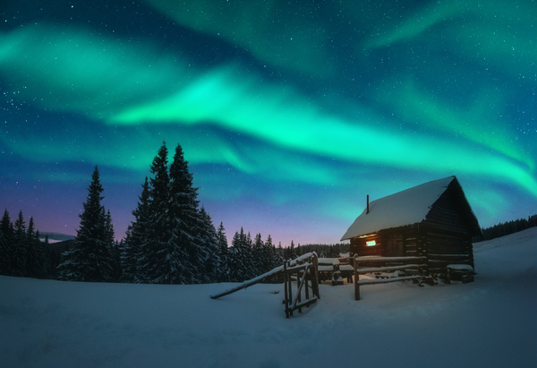 Fantastic winter landscape with wooden house with light in window in snowy mountains and northen light in night sky. Christmas holiday and winter vacations